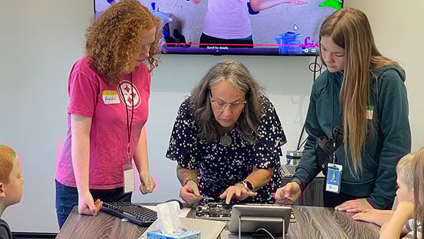 Woman taking apart a laptop with two female students observing.