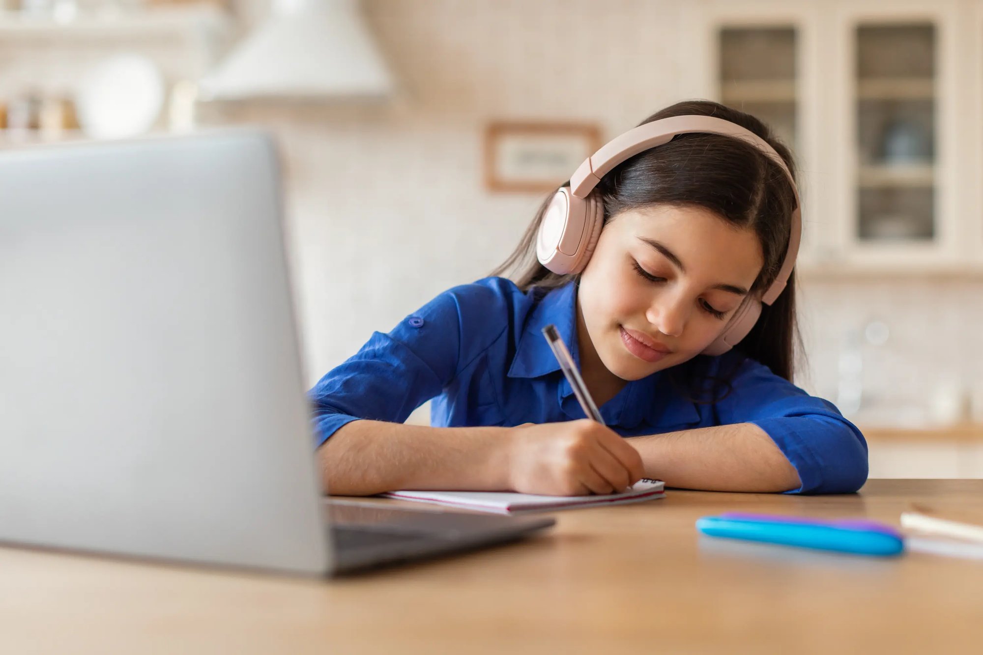 young girl wearing headphones doing homework while near a laptop