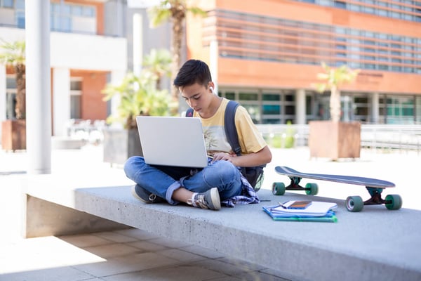 young male sitting outside looking at a laptop near a skateboard